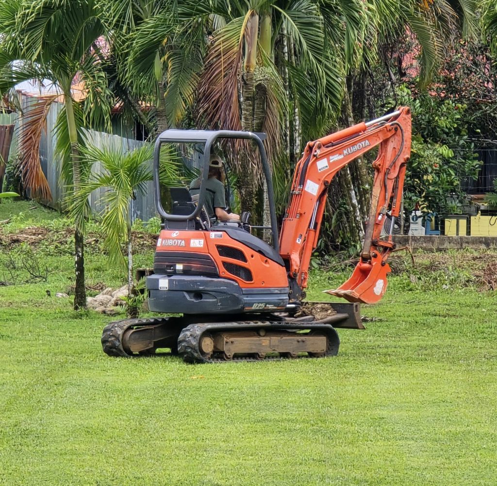 A small Kubota excavator in a lush green field, surrounded by palm trees, with an operator seated inside.