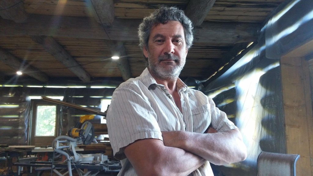 A man, Bob Gordon, is wearing a collared shirt and standing in a workshop environment with crossed arms and a slight smile.